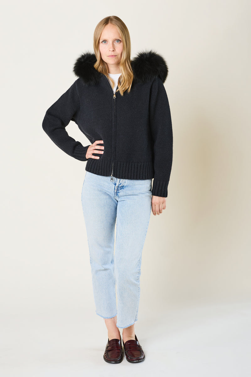 Woman wearing a black jacket with fur hood, light blue jeans, and brown shoes on a white background