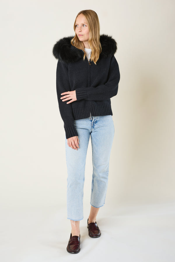 Woman wearing a black sweater with a fur-lined hood, light blue jeans, and dark shoes on a white background
