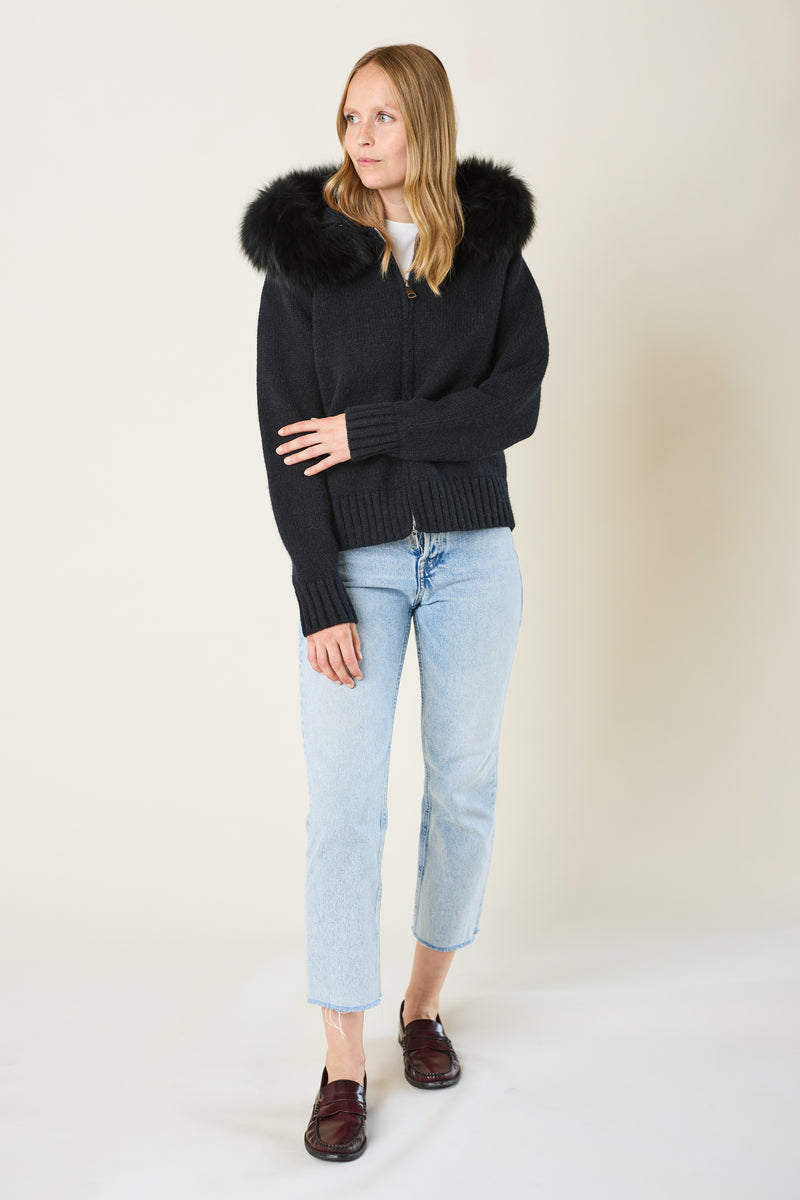 Woman wearing a black sweater with a fur-lined hood, light blue jeans, and dark shoes on a white background