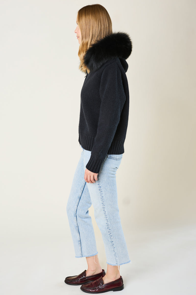 Woman wearing a black hoodie with fur hood and light blue jeans on a plain background
