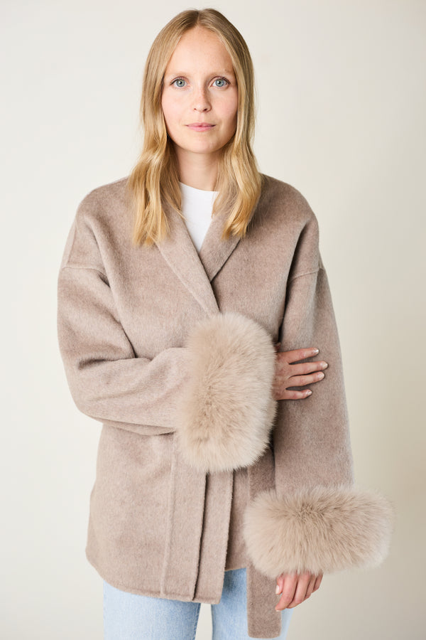 Woman wearing a beige coat with fur cuffs on a plain background