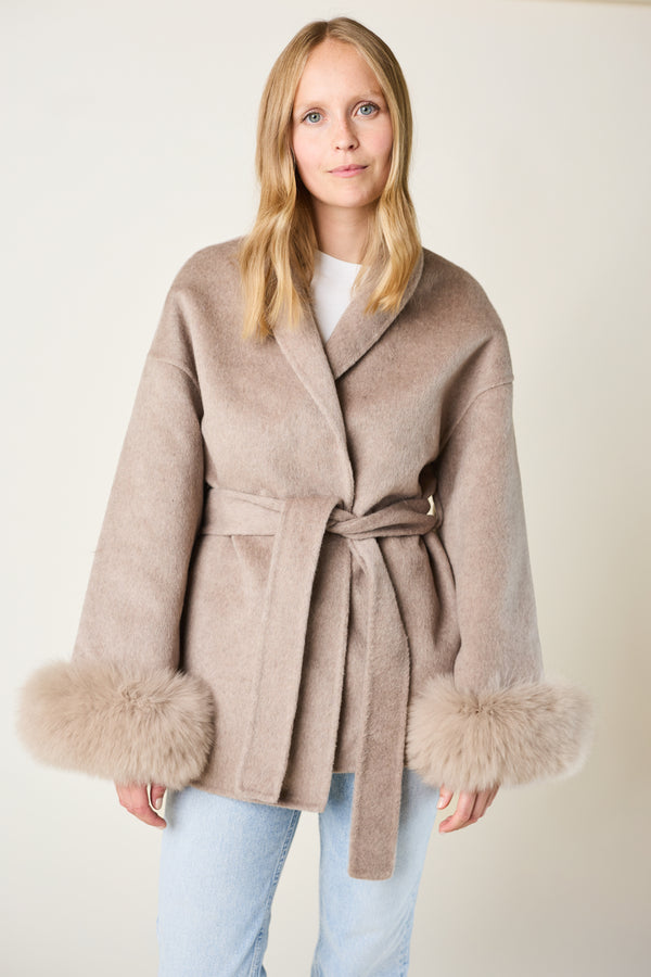 Woman wearing a beige coat with fur cuffs on a plain background