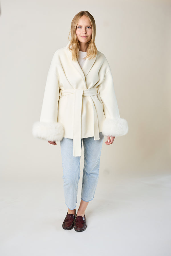 Woman wearing a cream-colored coat with fur trim on a plain background