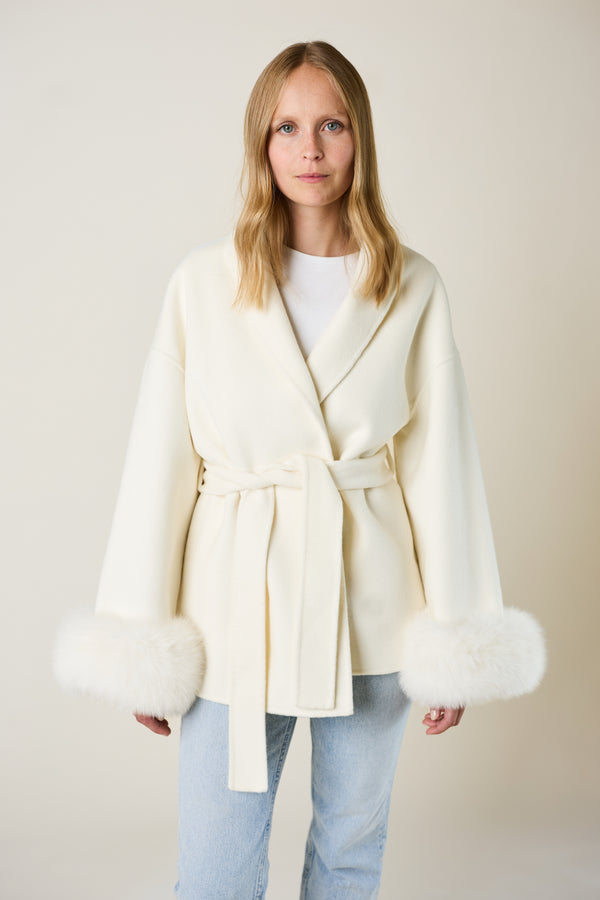 Woman wearing a cream-colored coat with fur trim on a plain background