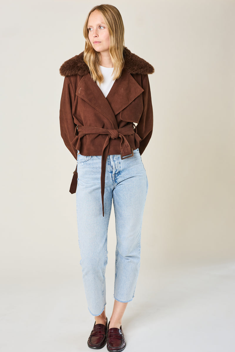 Woman wearing a brown coat with fur collar, white shirt, light blue jeans, and brown shoes on a plain background