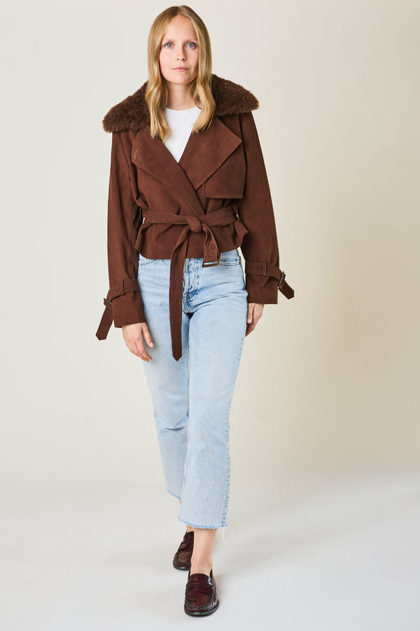 Woman wearing a brown coat with a fur collar, white shirt, and light blue jeans on a plain background