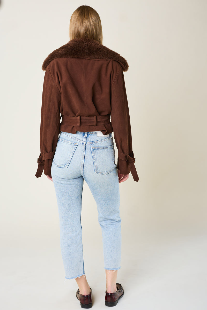 Person wearing a brown jacket with fur collar and light blue jeans on a plain background