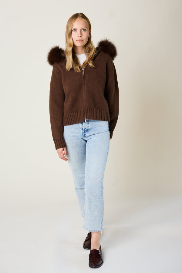 Woman wearing a brown sweater with fur hood and light blue jeans on a plain background