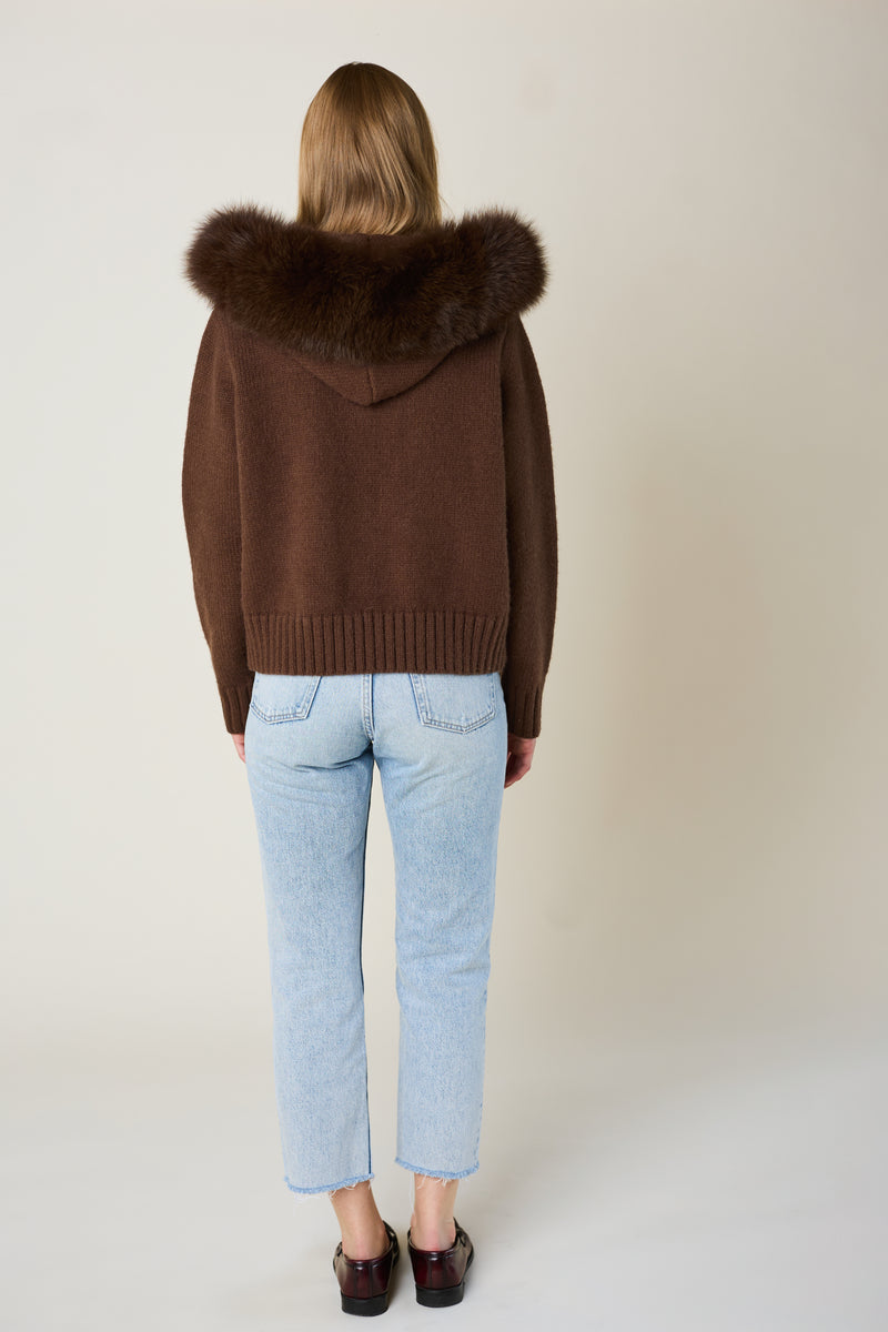 Person wearing a brown sweater with a fur hood and light blue jeans on a plain background