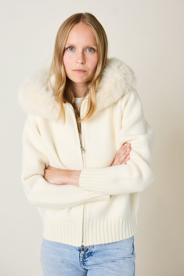 Woman wearing a cream-colored zip-up hoodie with fox fur hood against a plain background