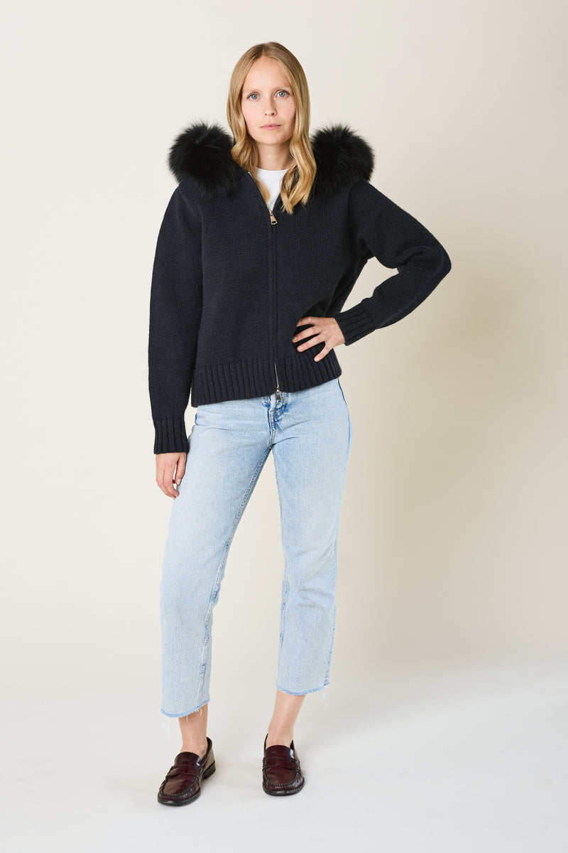 Woman wearing a black cardigan with fur trim, light blue jeans, and brown shoes on a white background